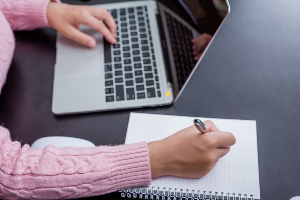 A female hospitality professional writing down content with her right hand in a notepad while using her left hand for her computer