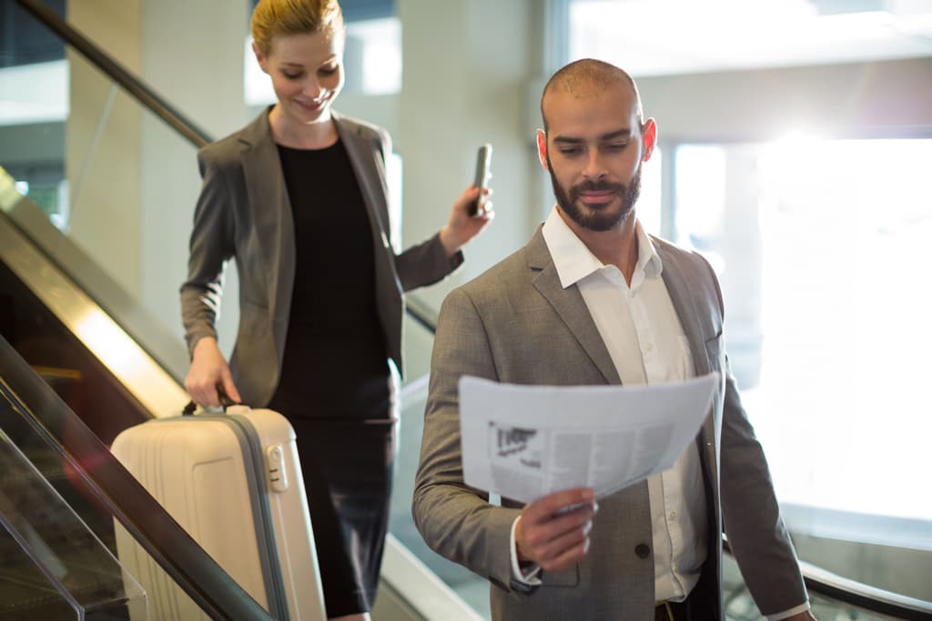 A businessman comes down a staircase wearing a sports jacket and reading some printed material while a female businesswoman follows behind the stairs while carrying a piece of luggage