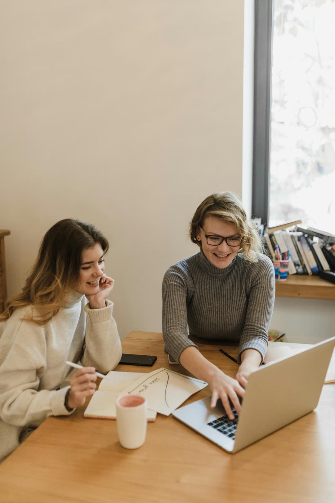 Two young women smiling and collaborating on a laptop and notebook at a wooden table in a brightly lit room.