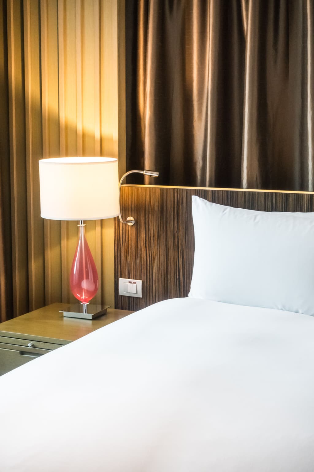 A hotel room with a bright lamp with a red base and a bed with white sheets