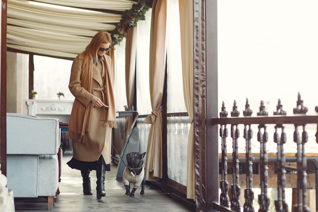 A woman walks with a dog on a leash in the outside porch of a property while wearing a trenchcoat