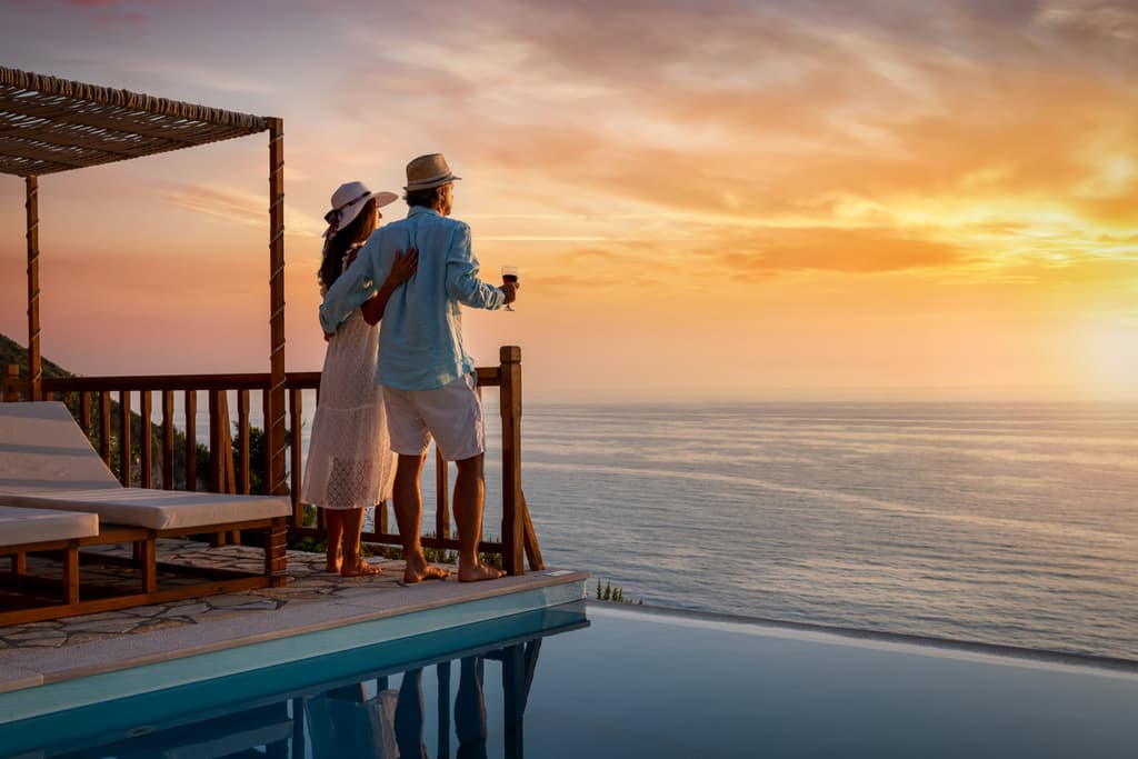 a couple from behind, standing on a stone patio by an infinity pool, looking out at a sunset over the ocean.